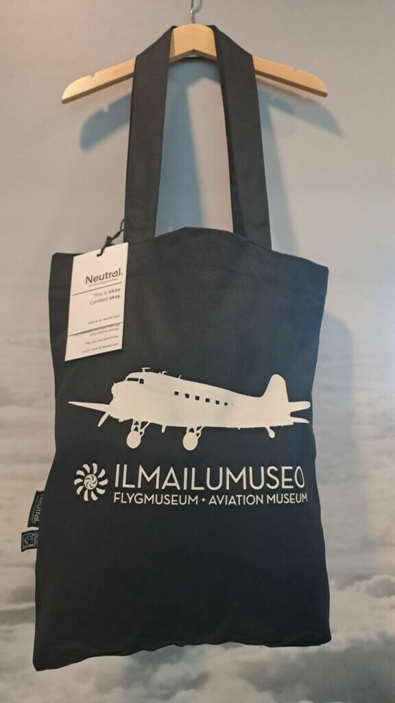 Ilmailumuseon musta kangaskassi henkarissa, näkyvissä lappu kankaan sertifikaateista.