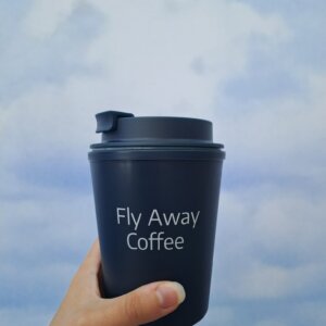 Henkilön kädessä tummansininen takeway-muki, jossa teksti "Fly Away Coffee".