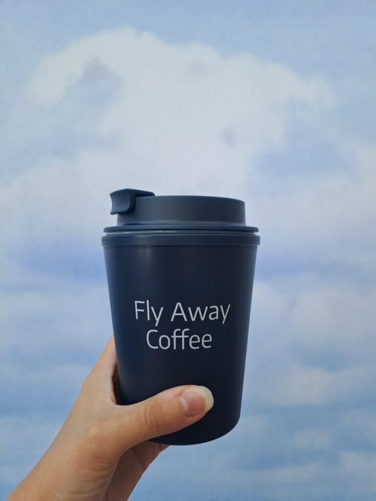 Henkilön kädessä tummansininen takeway-muki, jossa teksti "Fly Away Coffee".