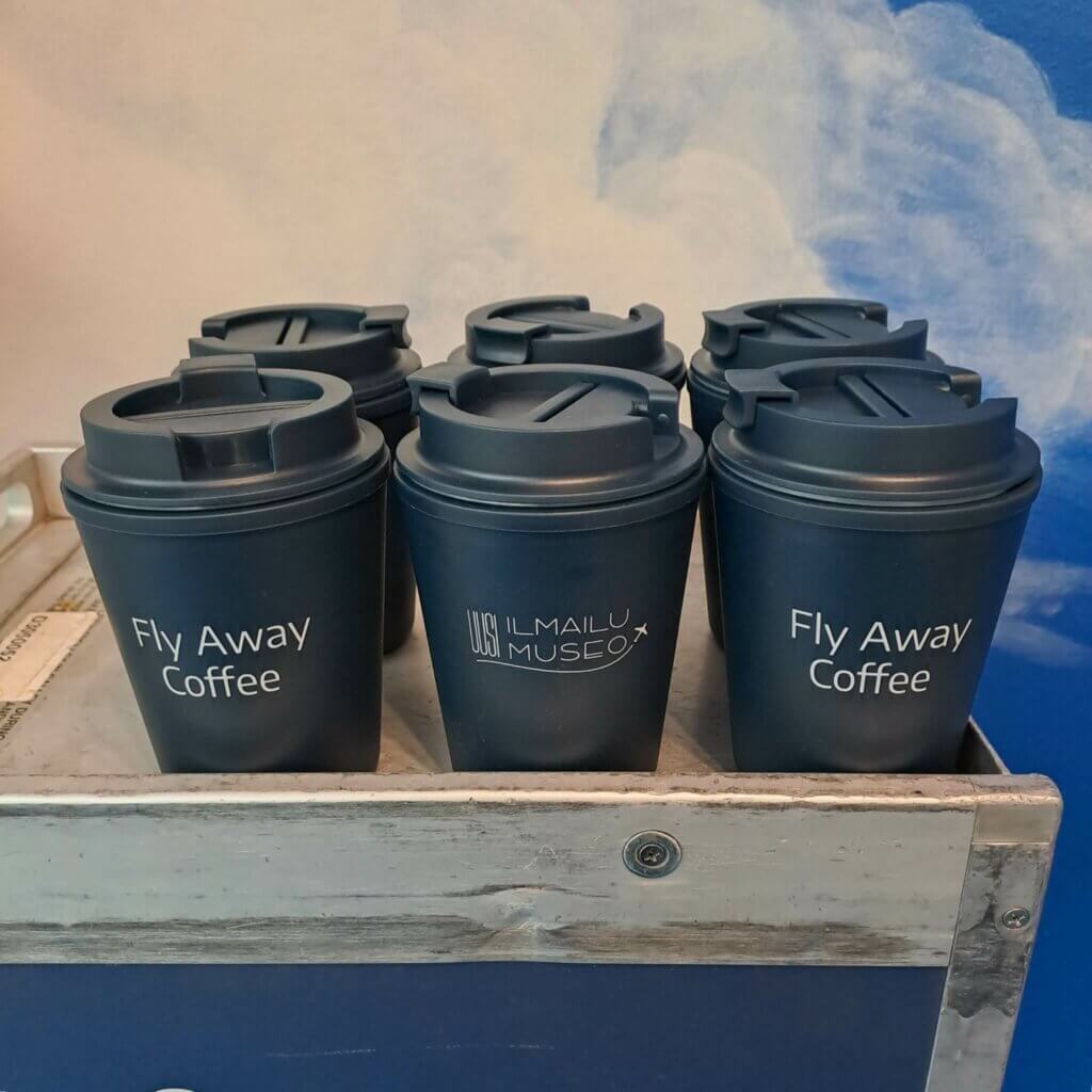 Tarjoilukärryssä useita sinisiä takeaway-mukeja, osassa näkyvissä logo, osassa teksti "Fly Away Coffee".