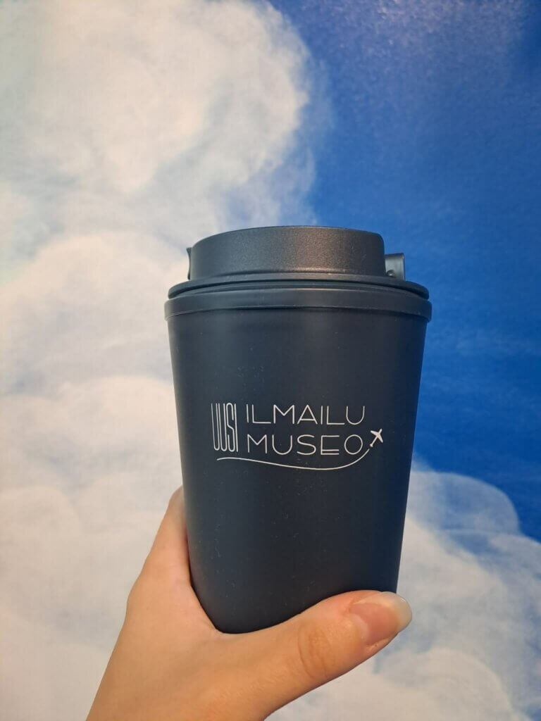 Henkilön kädessä tummansininen takeaway-muki, jossa Uusi Ilmailumuseo -logo.