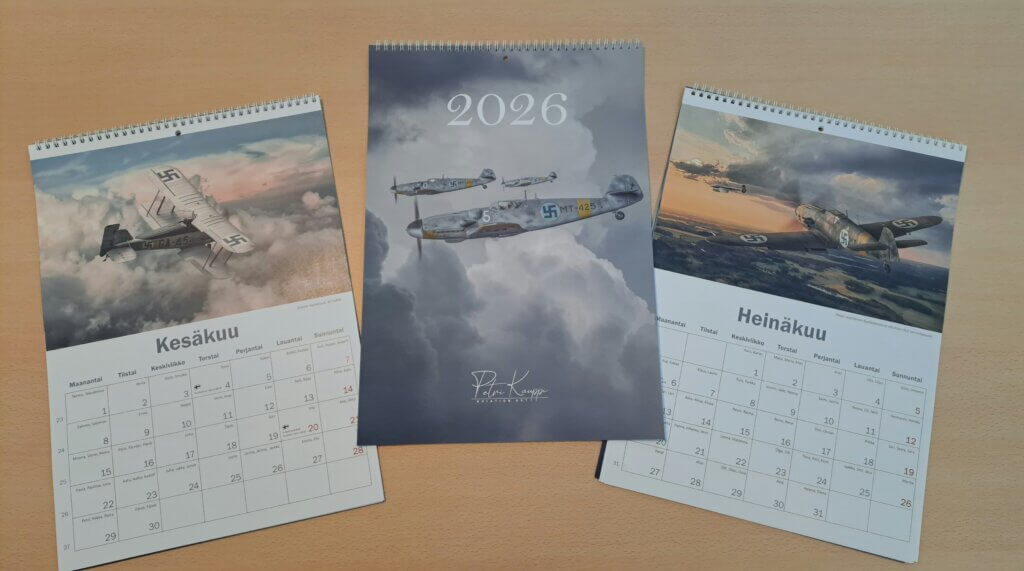 Kolme ilmailutaidekalenteria pöydällä, näkyvissä kansi sekä kaksi kuukausinäkymää.