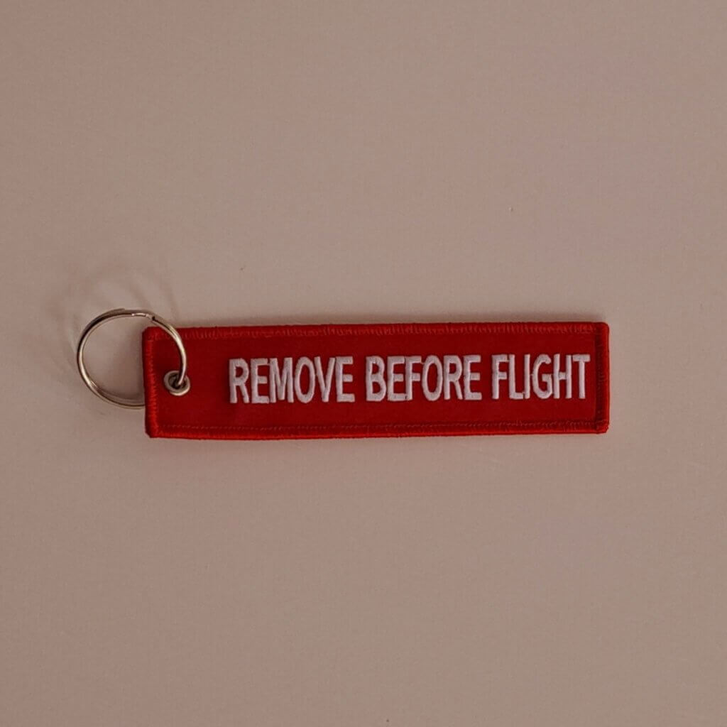 Punainen kangasavaimenperä, johon brodeerattu valkoisella "Remove before flight".