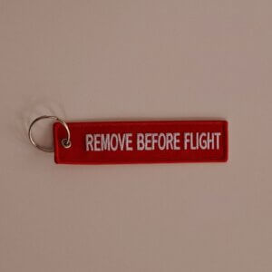 Punainen kangasavaimenperä, johon brodeerattu valkoisella "Remove before flight".