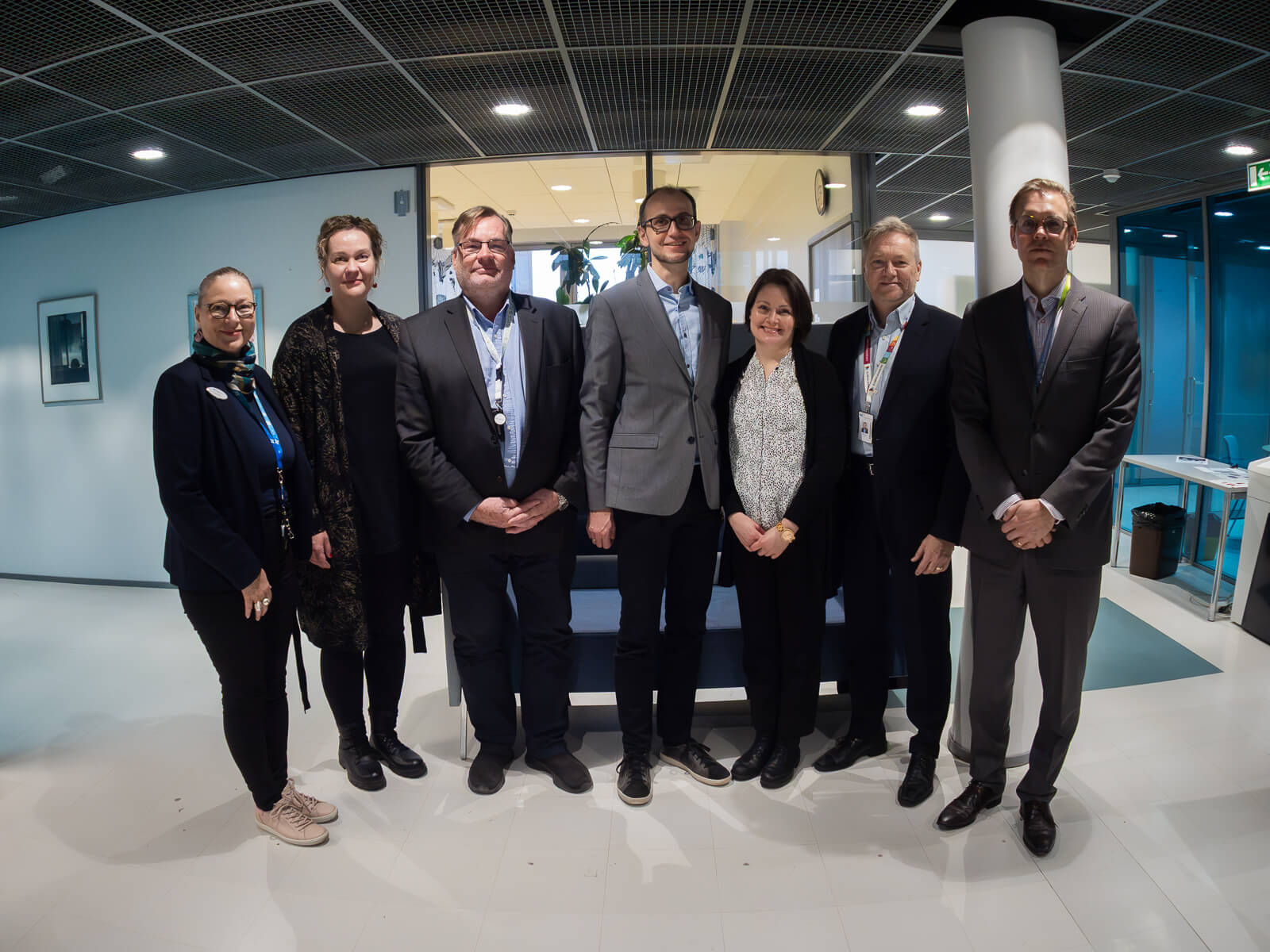 Ryhmänmuotoinen yhteiskuva seitsemästä henkilöstä modernissa toimistotilassa. Taustalla näkyy lasiseiniä, valaistuja toimistohuoneita ja kattoon asennettuja säleikkövalaisimia.