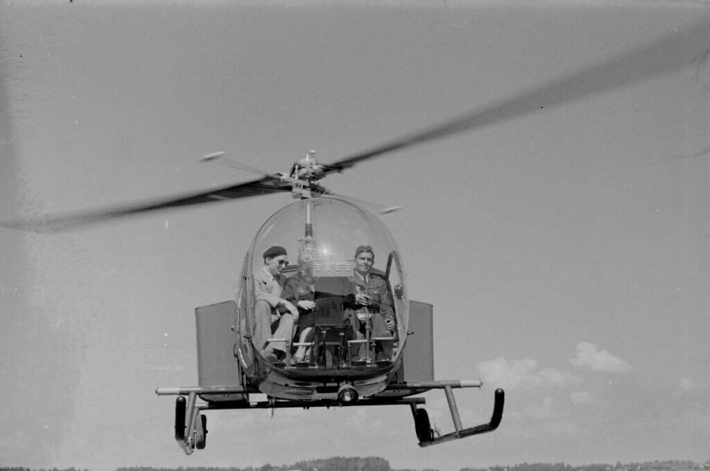 Kuvassa pieni helikopteri ilmassa edestä päin nähtynä. Helikopterin ohjaamossa on kolme ihmistä.