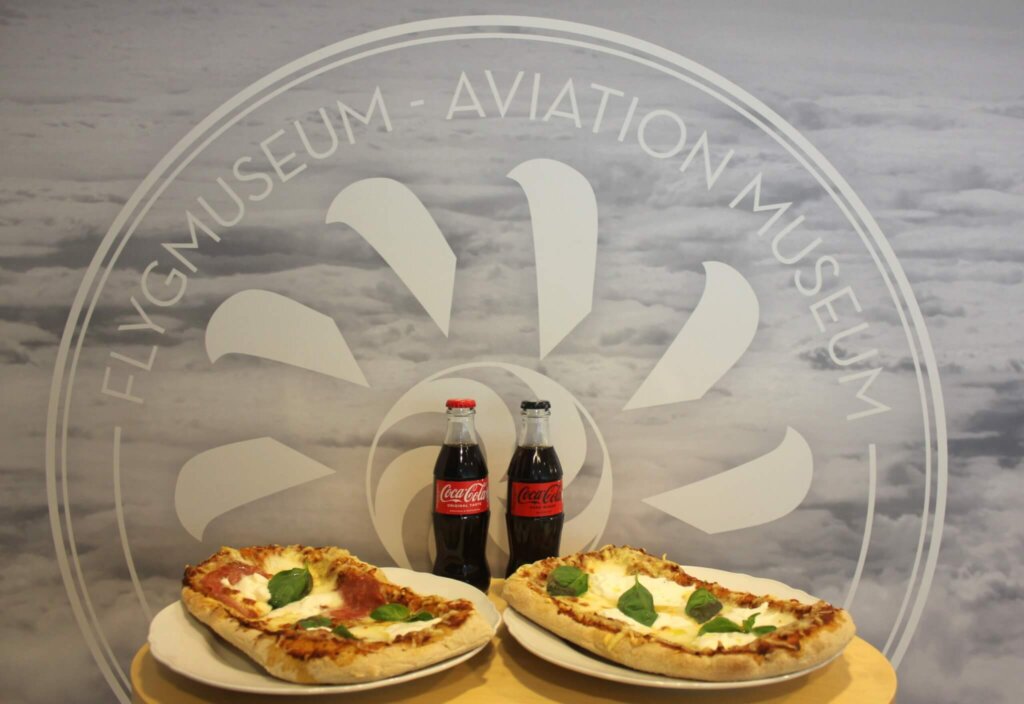 Pöydällä kaksi pizzaa ja kaksi Coca-Colaa, taustalla vaalea pilvimaisema ja Suomen Ilmailumuseon logo.