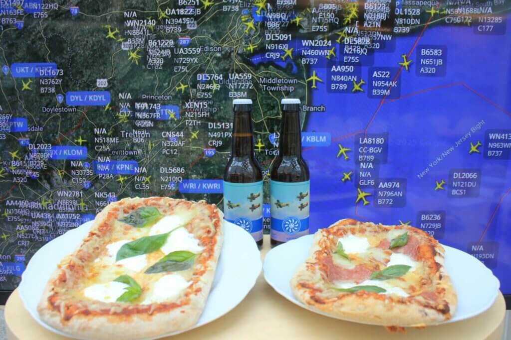 Kaksi pizzaa ja kaksi pulloa olutta pöydällä, taustalla Flight Radarin näkymä New York Citysta.