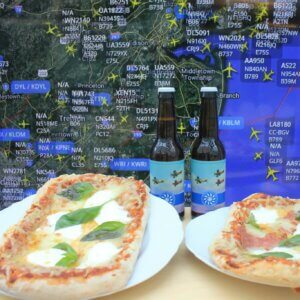 Kaksi pizzaa ja kaksi pulloa olutta pöydällä, taustalla Flight Radarin näkymä New York Citysta.