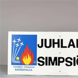 Kyltti, jossa värikäs tarrakuva tekstillä "Lentäjien juhannus Kauhavalla". Lisäksi kyltissä lukee "Juhlaradio Simpsiö 96.9".
