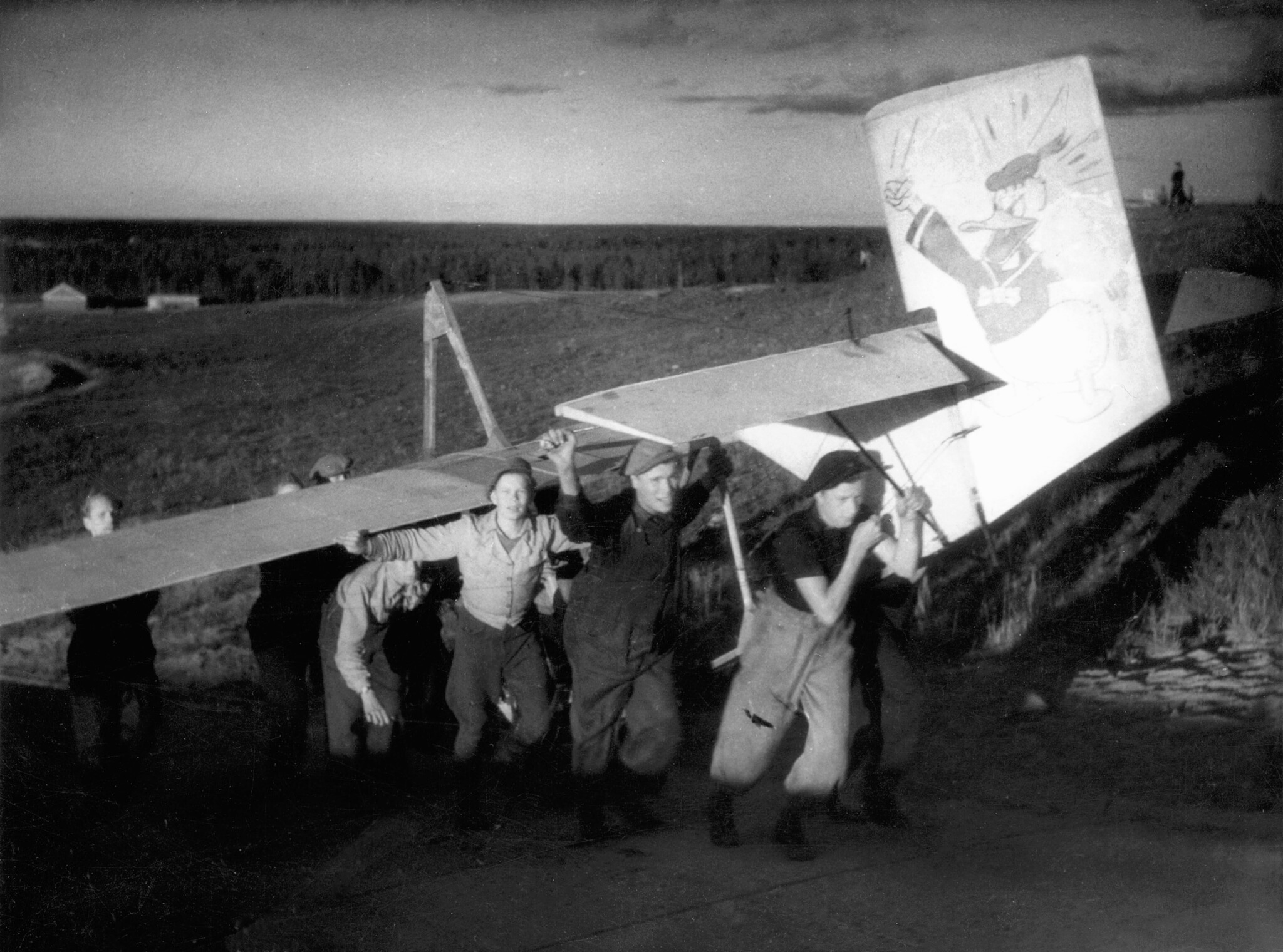 Kuvassa joukko nuoria miehiä kantamassa purjelentokonetta ylös mäkeä. Lentokoneen pystyvakaajassa on maalattuna sarjakuvahahmo.