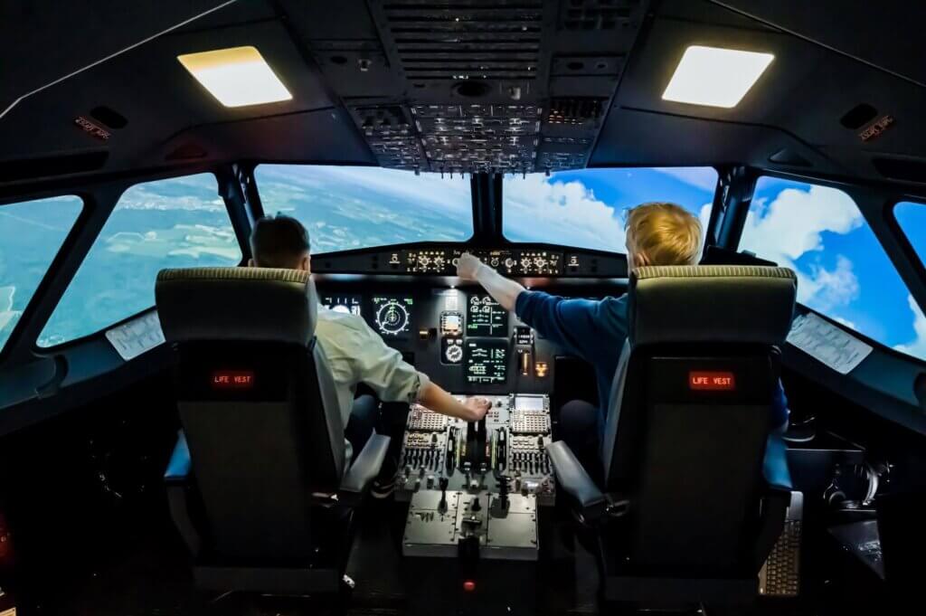 Kaksi nuorta aikuista lentämässä Airbus A320-simulaattorilla, joka kaartaa vasemmalle. "Koneen" vasemmalla näkyy maata ja oikealla taivasta.
