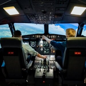 Kaksi nuorta aikuista lentämässä Airbus A320-simulaattorilla, joka kaartaa vasemmalle. "Koneen" vasemmalla näkyy maata ja oikealla taivasta.