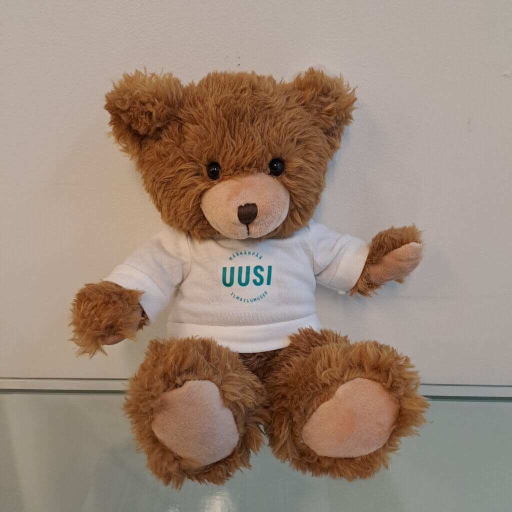 Ruskea pehmonalle, jolla on valkoinen t-paita vihreällä Uusi Ilmailumuseo -logolla.