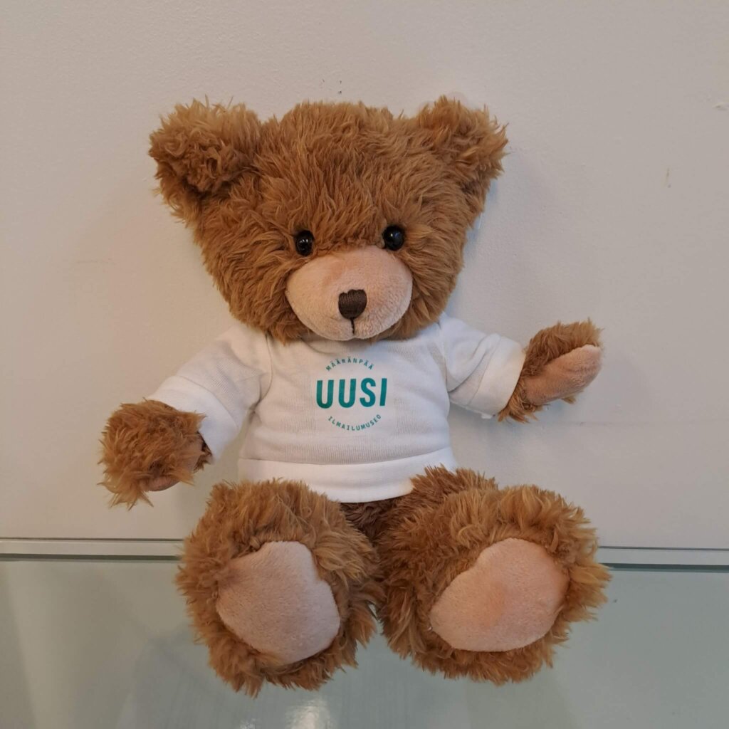 Ruskea pehmonalle, jolla on valkoinen t-paita vihreällä Uusi Ilmailumuseo -logolla.